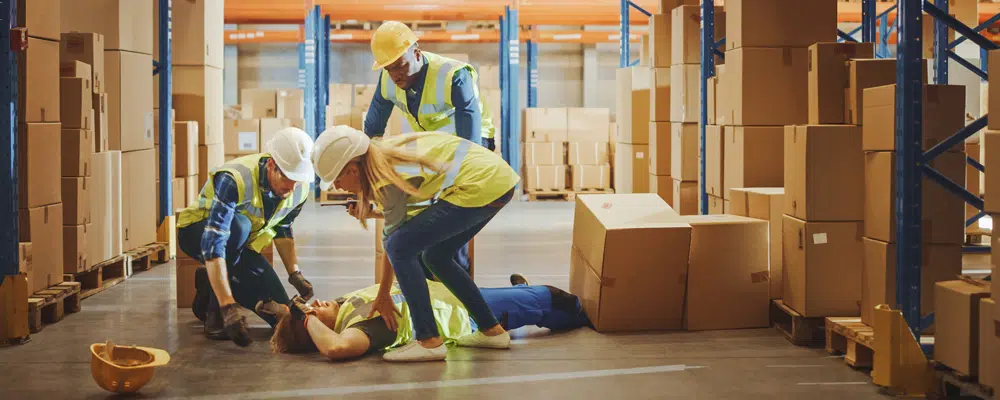 a group of people in a warehouse wearing protective gear (hard hat) helping a person on the floor how has been hit by some boxes.