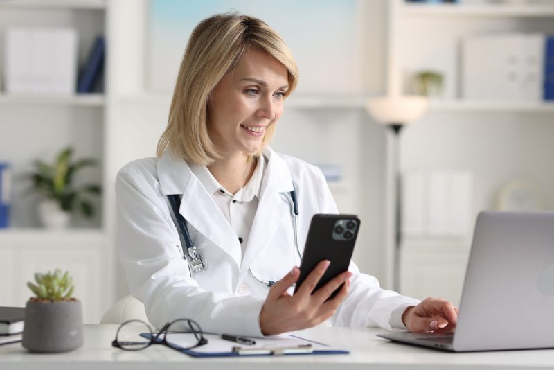 Smiling,Doctor,With,Smartphone,And,Laptop,Having,Online,Consultation,At